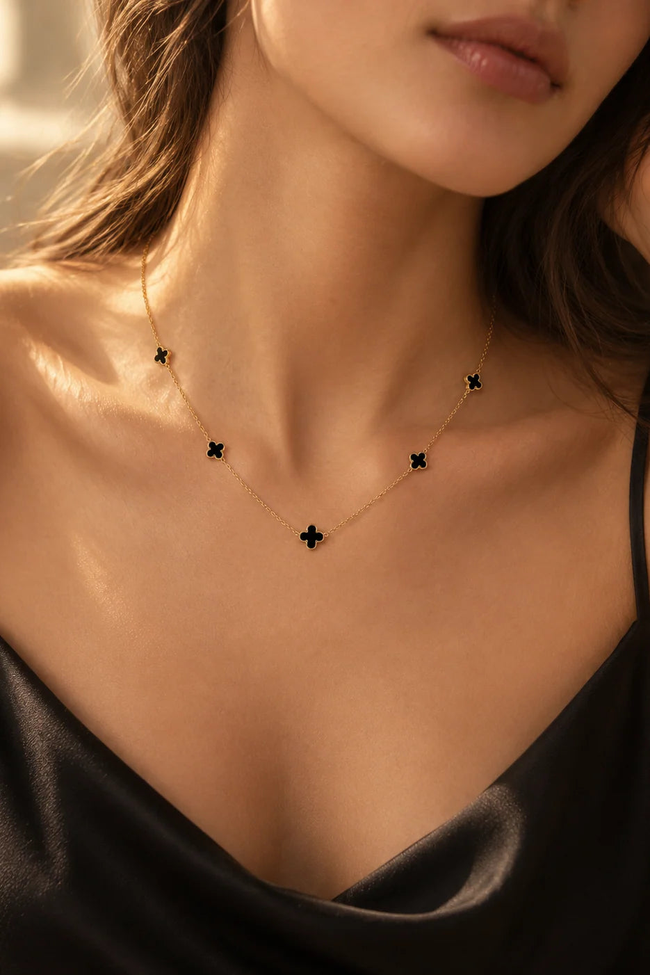 A close-up lifestyle shot of a model wearing a delicate gold chain necklace with five small black clover-shaped charms, styled with a black satin top.