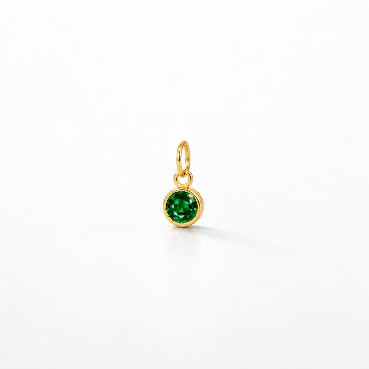 A small gold charm pendant featuring a round, faceted green gemstone in a bezel setting, suspended from a gold loop, shown on a clean white background.