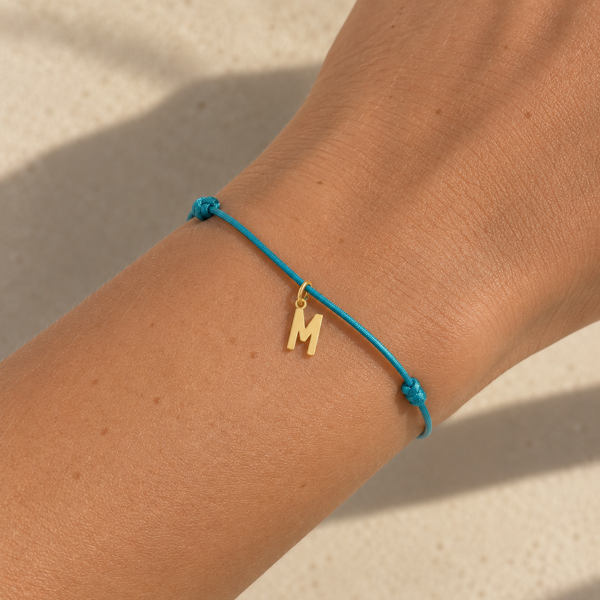 A turquoise blue cord bracelet with a small gold initial 'M' charm, worn on a person's wrist against a warm beige background.
