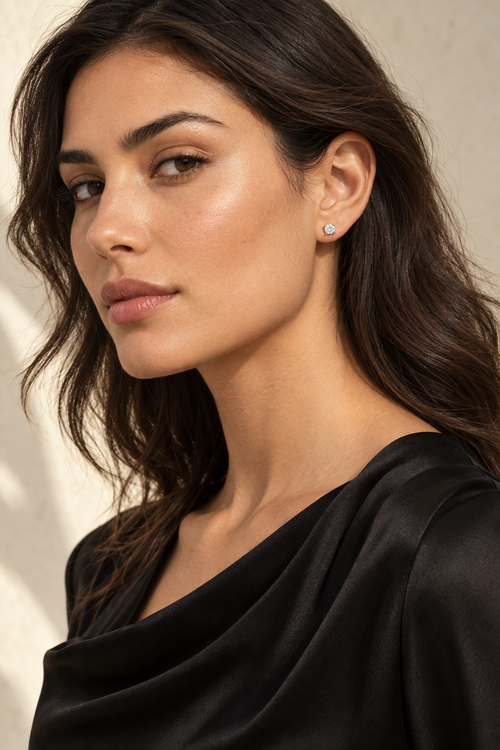 A close-up portrait of a woman with dark hair wearing a small silver stud earring with a clear gemstone, set against a soft beige background.