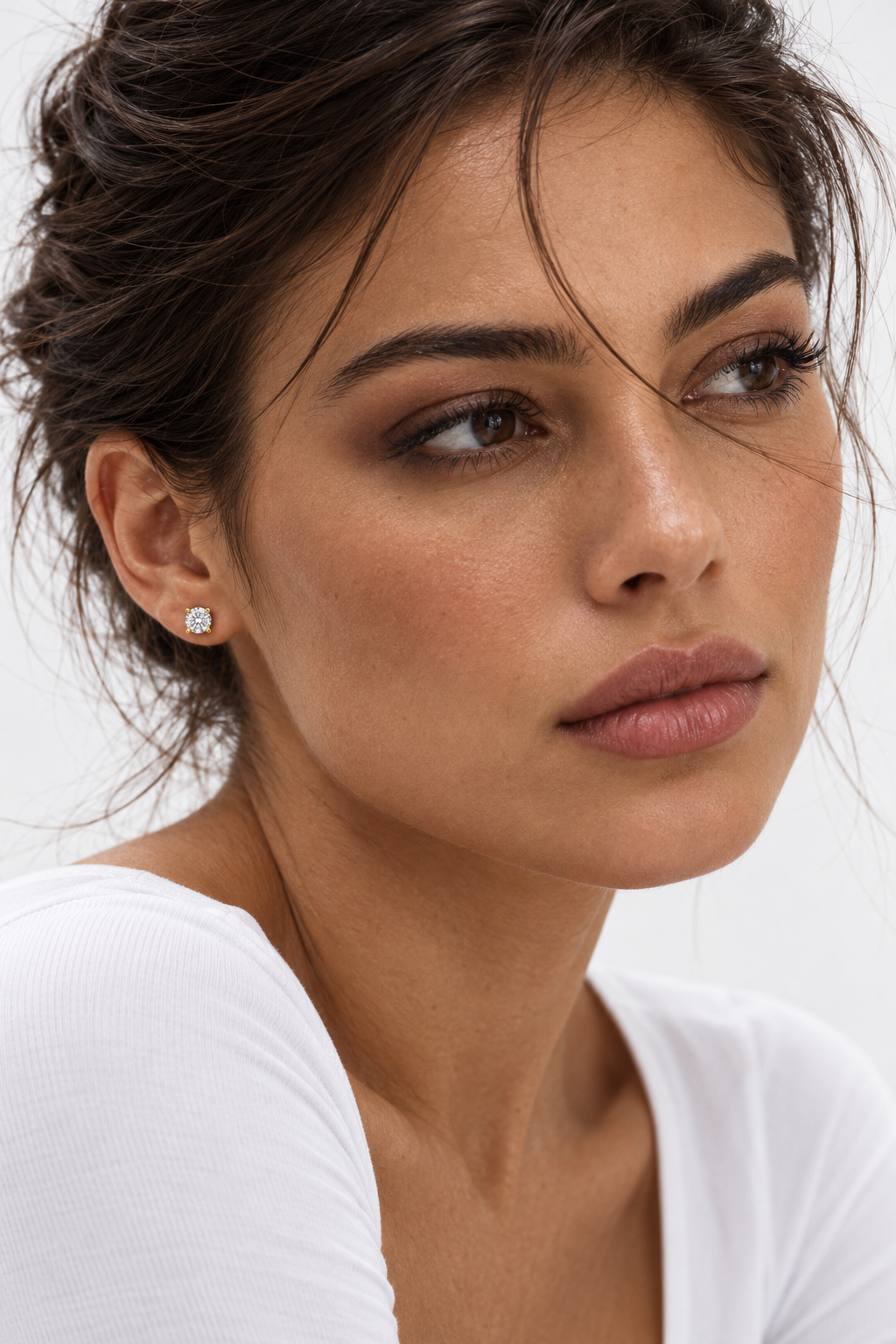 AI-generated woman with swept-up dark hair and a white top wearing a round clear CZ stud in a gold-plated four-claw setting.